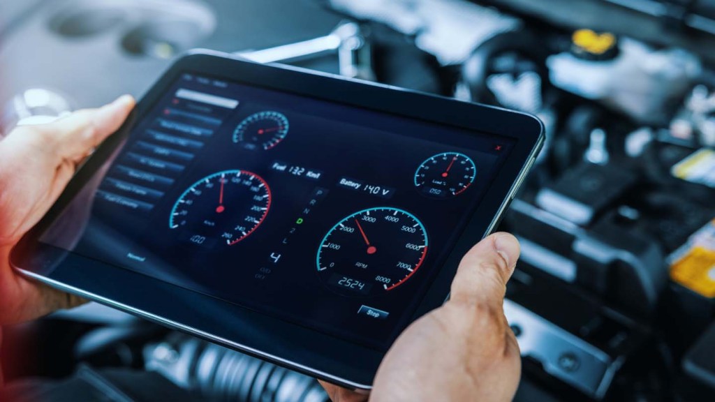 Hands hold a tablet over a car engine. The car's digital data is on the screen.