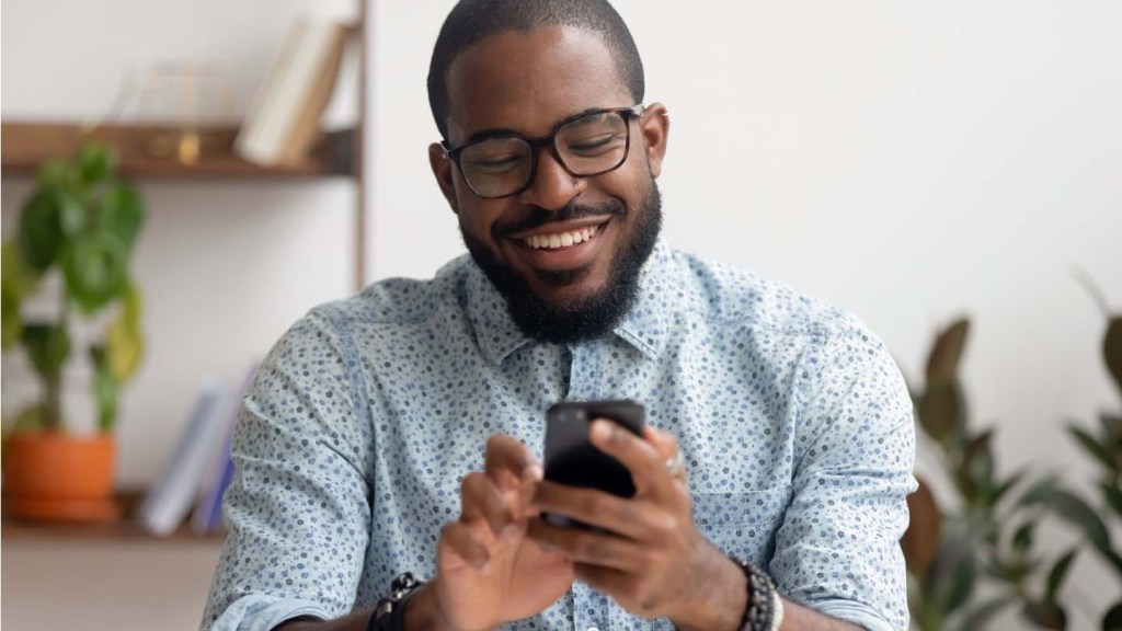 A smiling man with a beard and glasses holds his mobile device in front of him with two hands.
