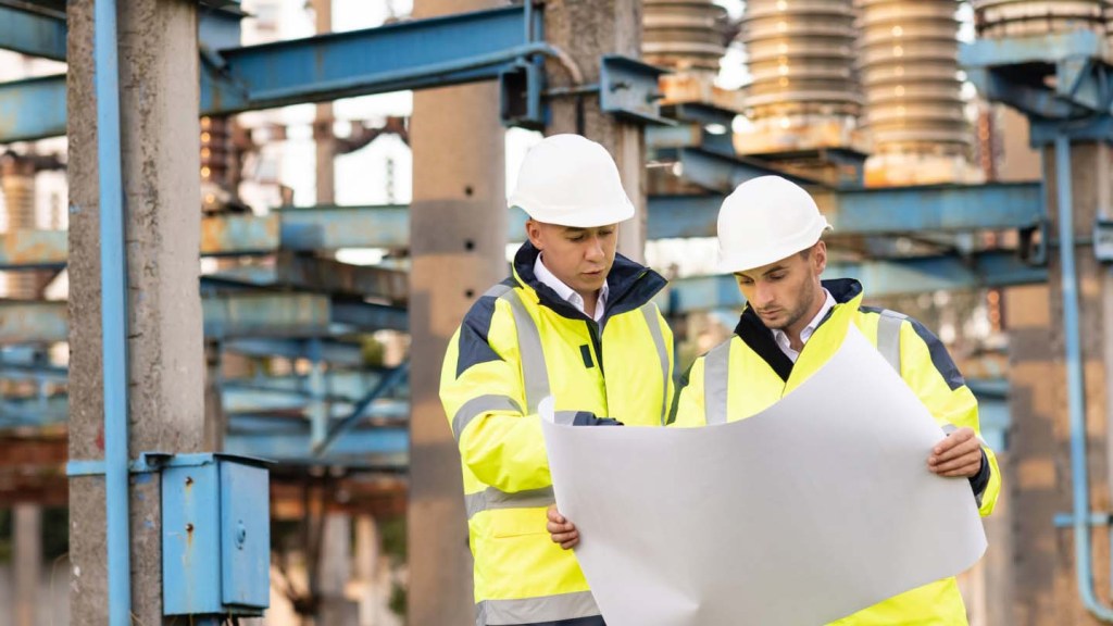 Two men in yellow jackets and hard hats look at a blueprint while at a construction site.
