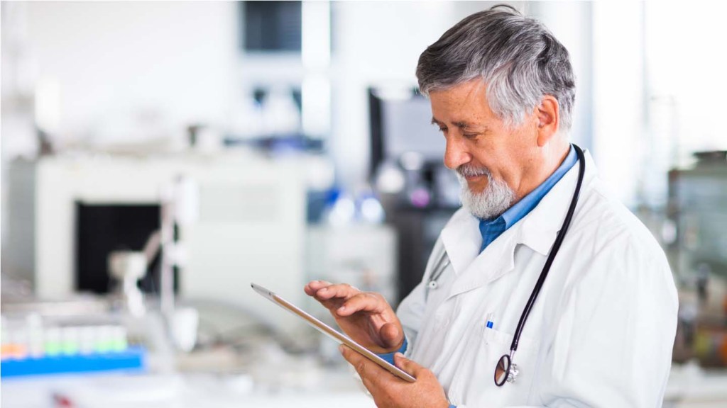 An older, male doctor is wearing a white lab coat and stethoscope in a medical lab. He looks down at his tablet.
