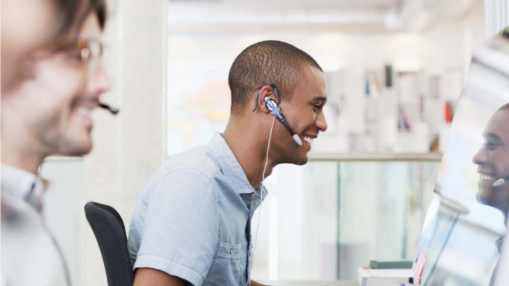 Two male customer support techs with headsets on smile as they look at their computer screens.