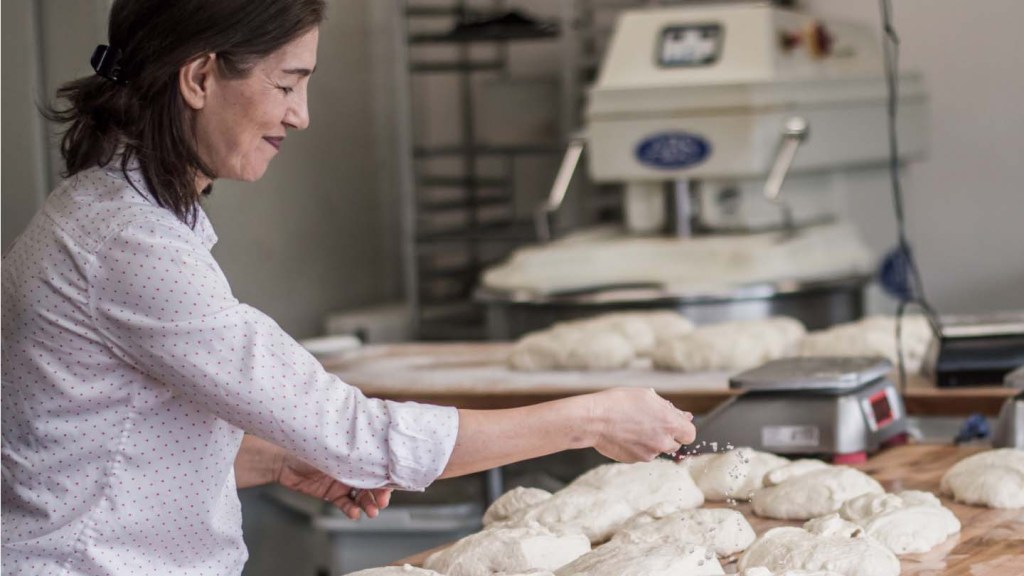 A woman spreads flour over bread dough about to be baked in an industrial kitchen.