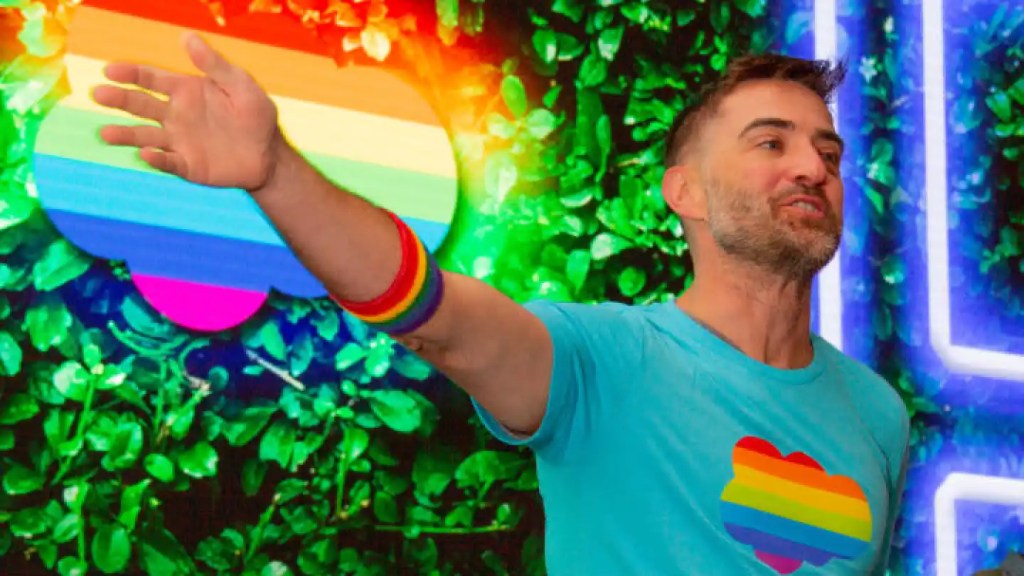 A man standing and smiling in front of a pride flag themed Salesforce logo