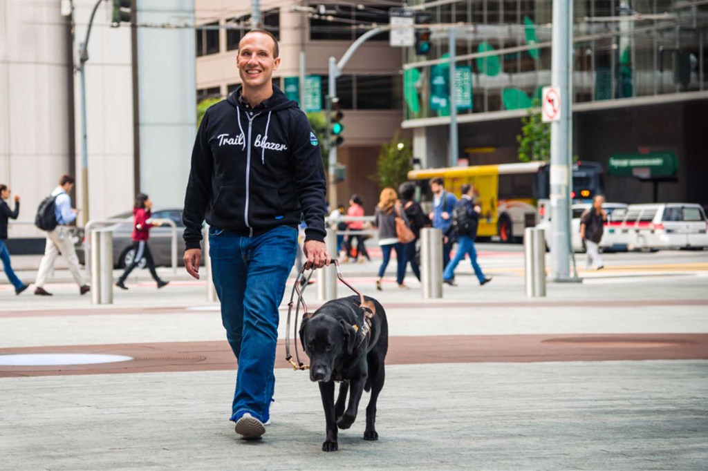 Man wearing Trailblazer hoodie crossing street witih guide dog.